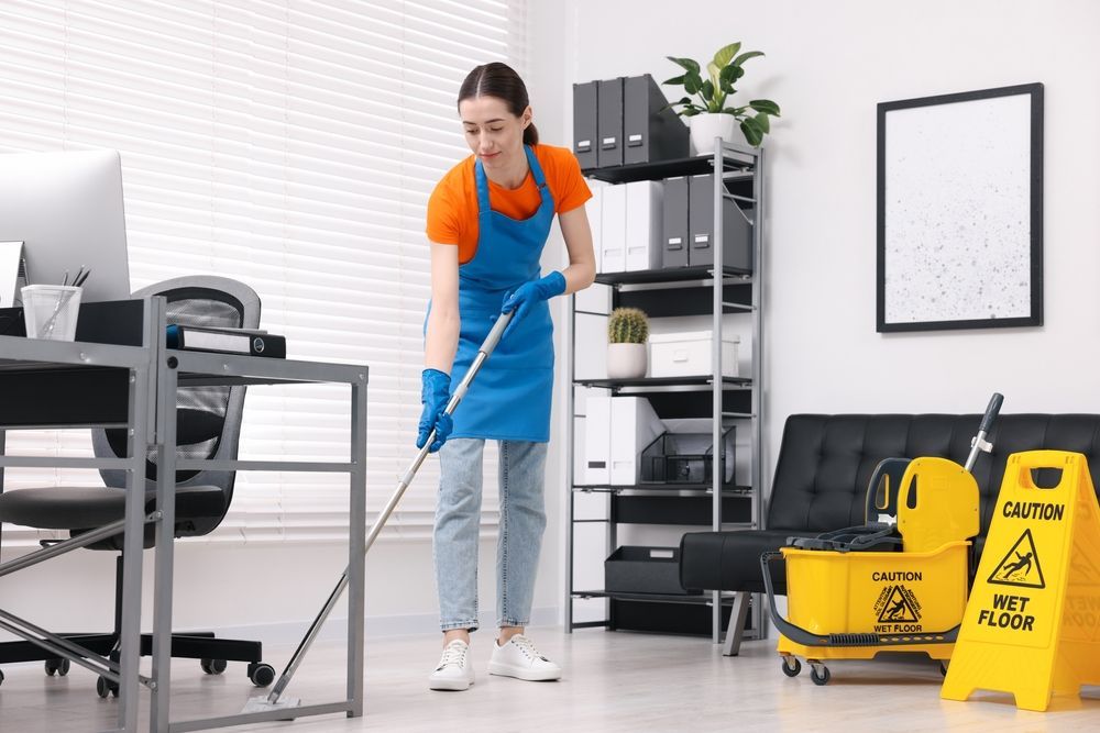 Person mopping an office floor; wearing an orange shirt, blue apron, and gloves. Yellow wet floor sign nearby.