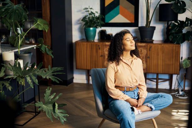 Woman sitting in a chair, eyes closed, surrounded by plants. Warm-toned room with wooden furniture and colorful art.