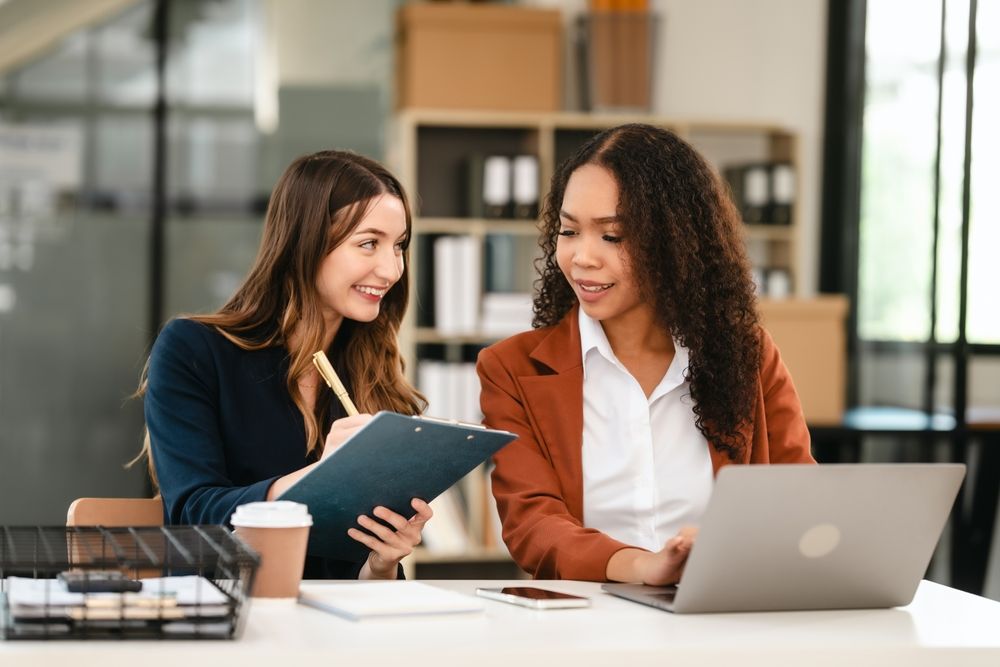 Two women working at a desk, one with a laptop, the other with a clipboard. Office setting, focused expressions.
