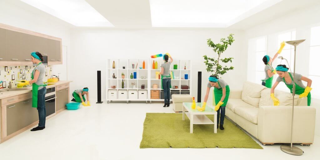 Multiple people in green aprons cleaning a bright, white living space, kitchen and couch.