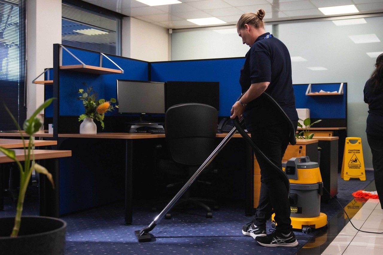 Person vacuuming the carpeted floor in an office cubicle, wearing a navy uniform.