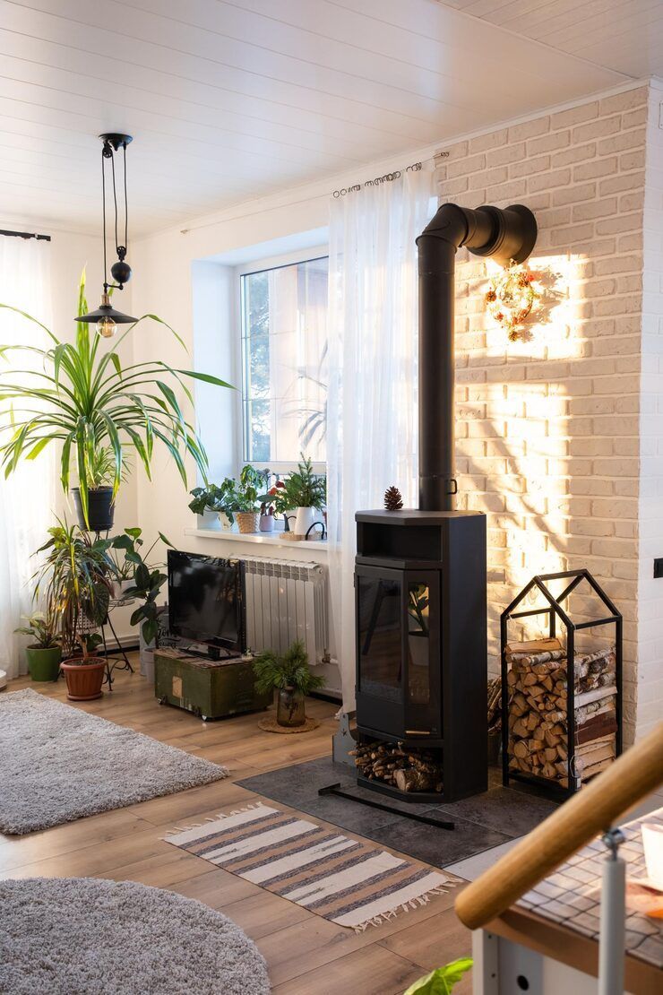 Cozy living room with a black wood-burning stove, plants, and a brick accent wall with sunlight.