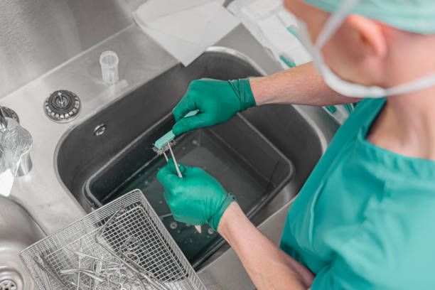 Person in green scrubs and gloves cleaning surgical tools in a stainless steel sink.