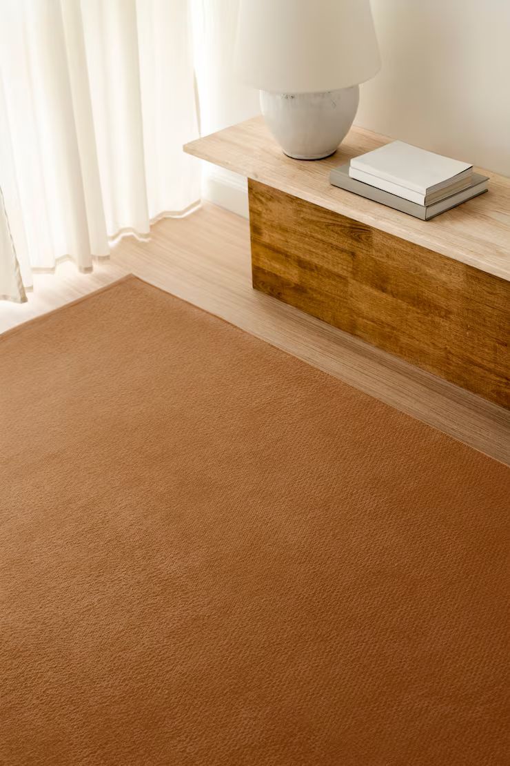 Brown rug on wooden floor, near a wooden console with a lamp and books.