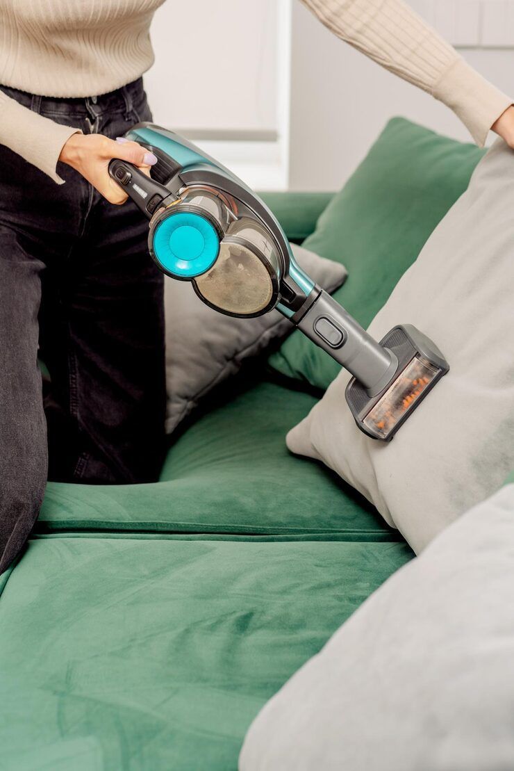 Person vacuuming a grey pillow on a green couch with a cordless vacuum cleaner.