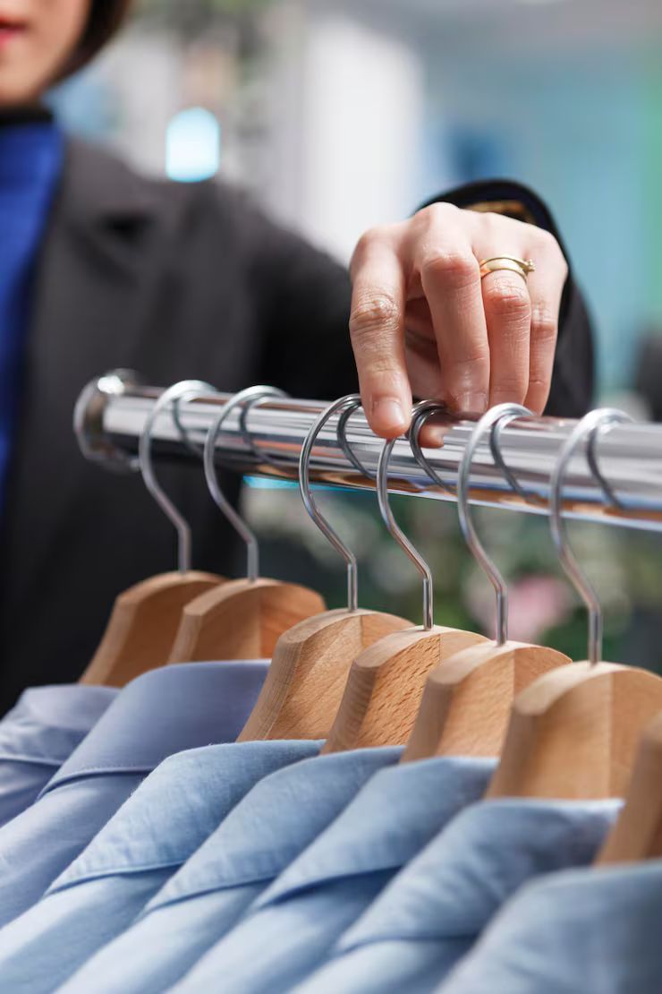 Person's hand reaching for a light blue shirt on a wooden hanger, part of a clothing rack in a store.