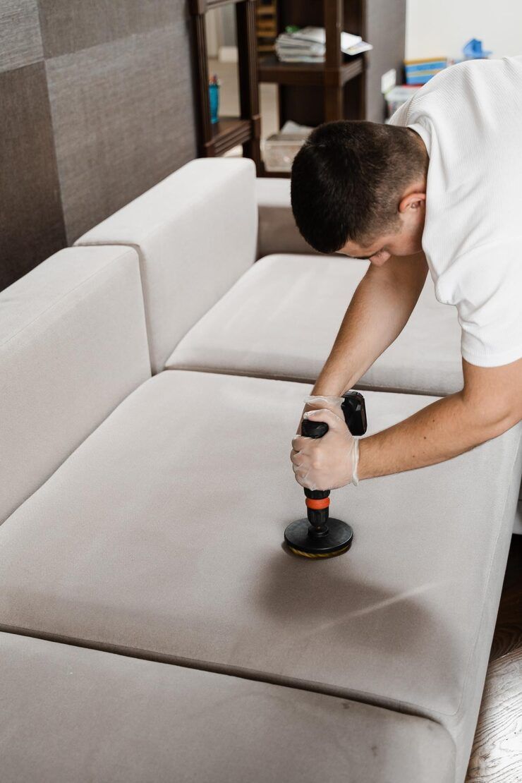 Person cleaning a light-colored sofa with a handheld machine indoors.
