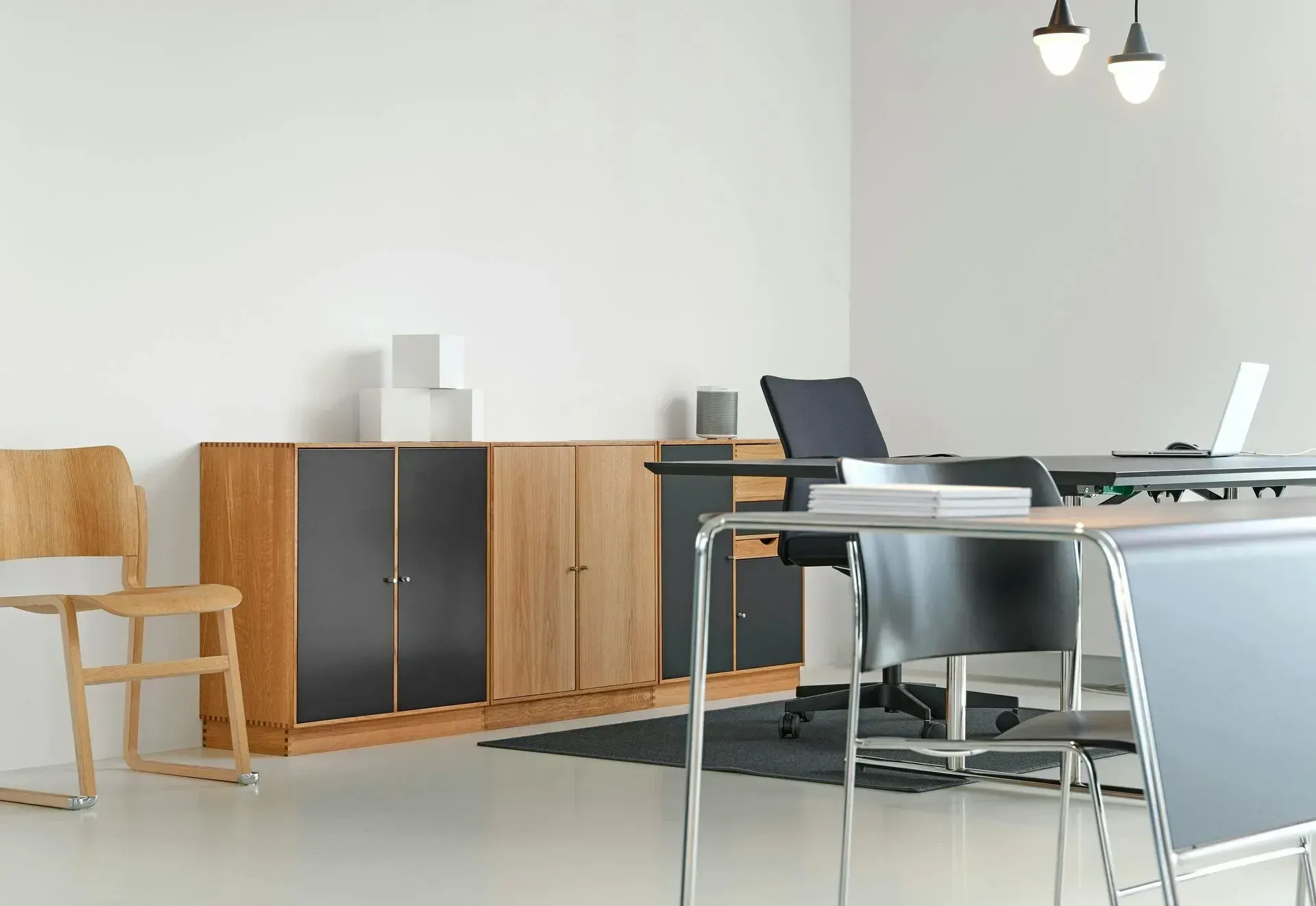 A minimalist office with a wood and black storage cabinet, a wooden chair, and desks with office chairs on a rug.