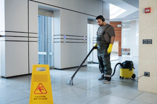 Custodian vacuuming floor, caution sign present, in an office or lobby setting.