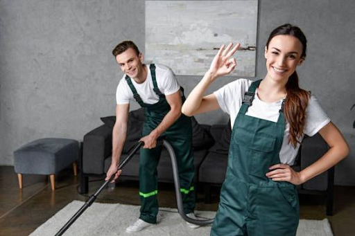 Woman in green overalls makes okay sign, smiling, while man vacuums a carpet in a living room.