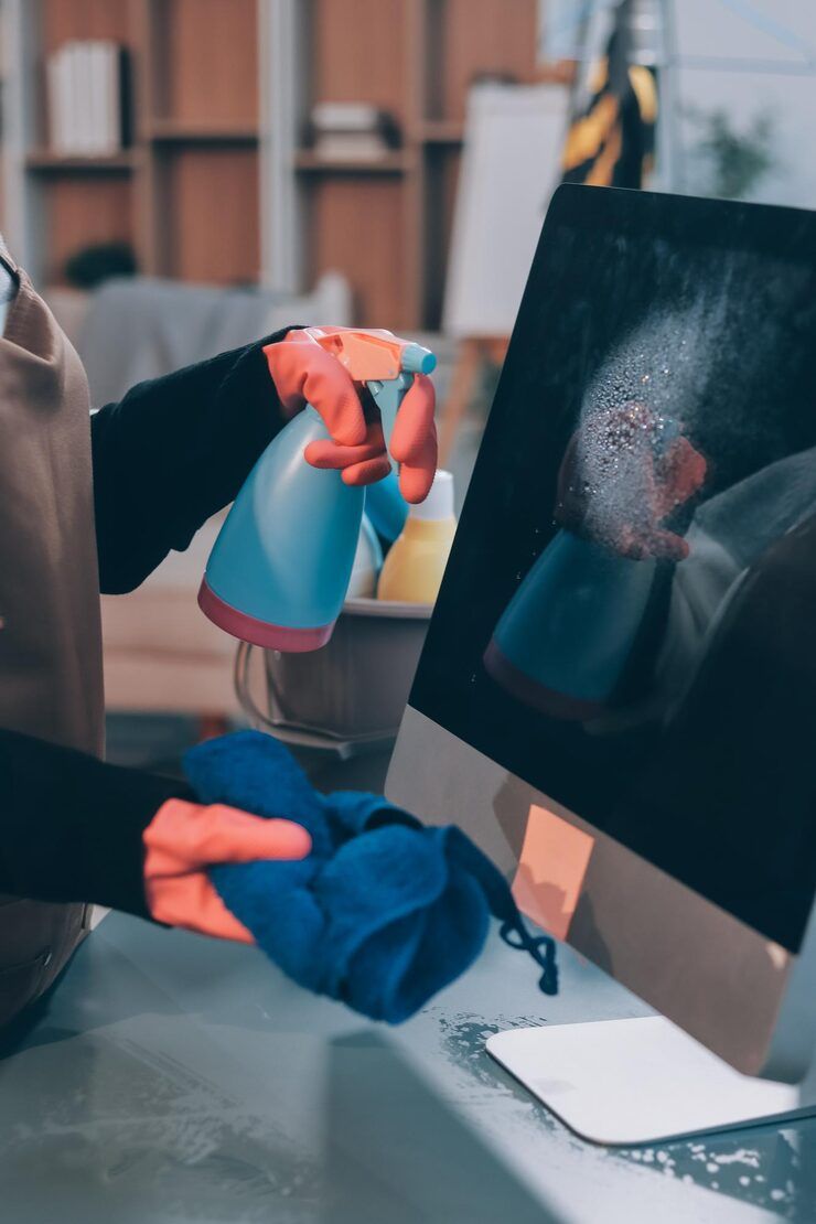 Person wearing gloves sprays cleaner on a computer screen and wipes it with a blue cloth.