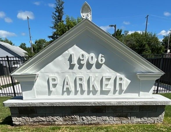 White sign with raised “1906 PARKER” lettering on a stone base outdoors under a blue sky.