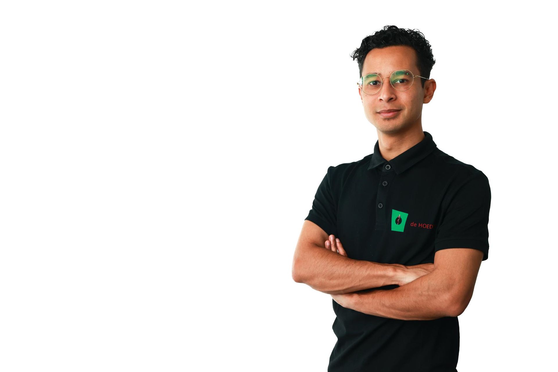 Een man met een bril en gekruiste armen, gekleed in een zwart poloshirt met een groen logo, staat voor een witte achtergrond.