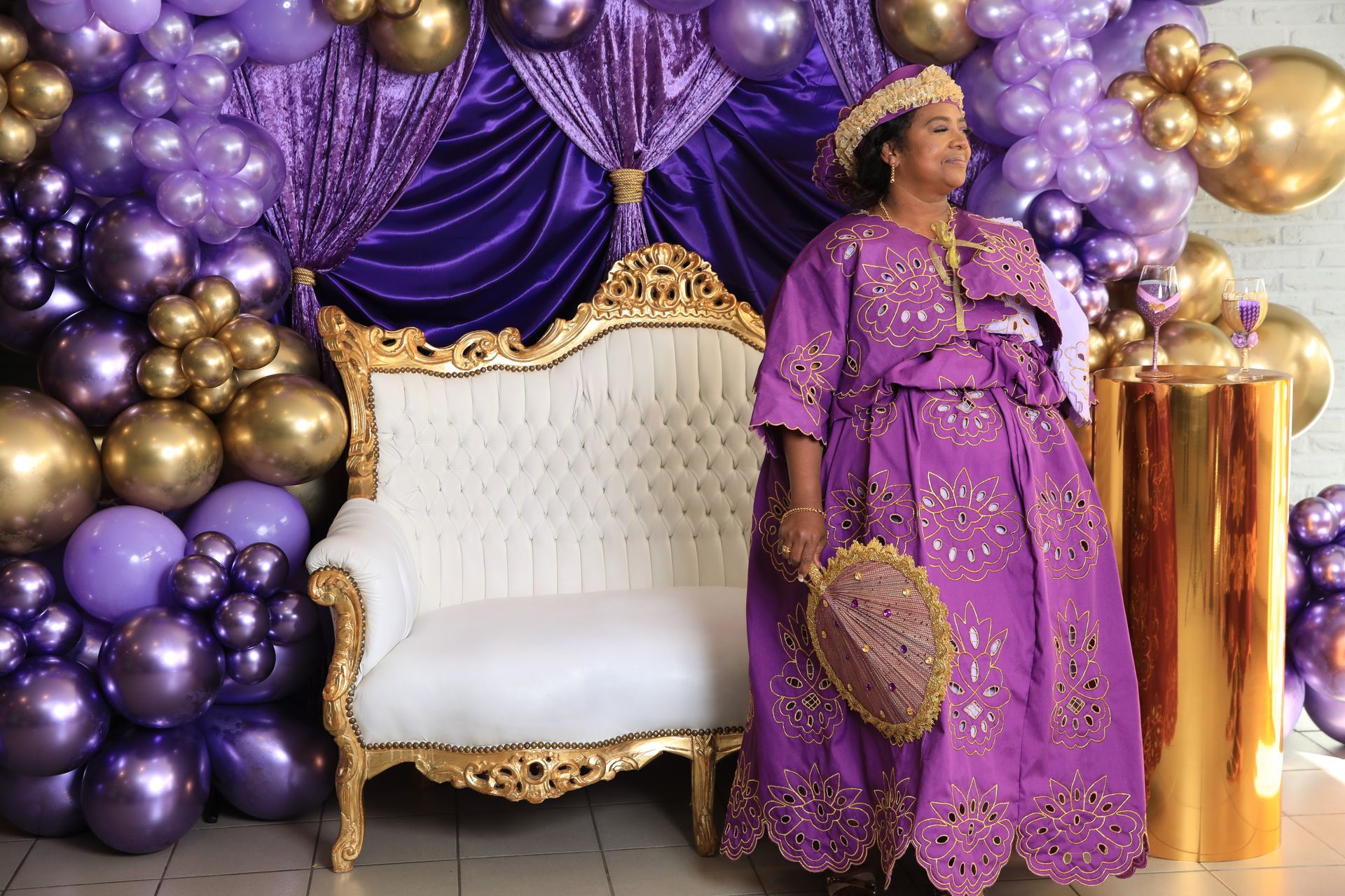 Een vrouw in een paarse jurk en met een kroon staat bij een vergulde stoel, voor een paarse en gouden ballonnenboog.
