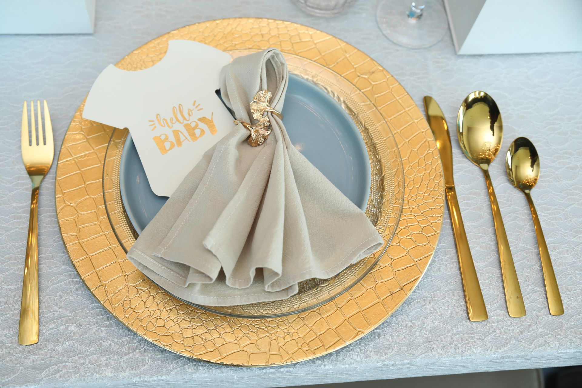 Tafeldekking met gouden bestek en onderbord, beige servet met gouden ring, kaartje met babythema, op een lichtblauw bord.