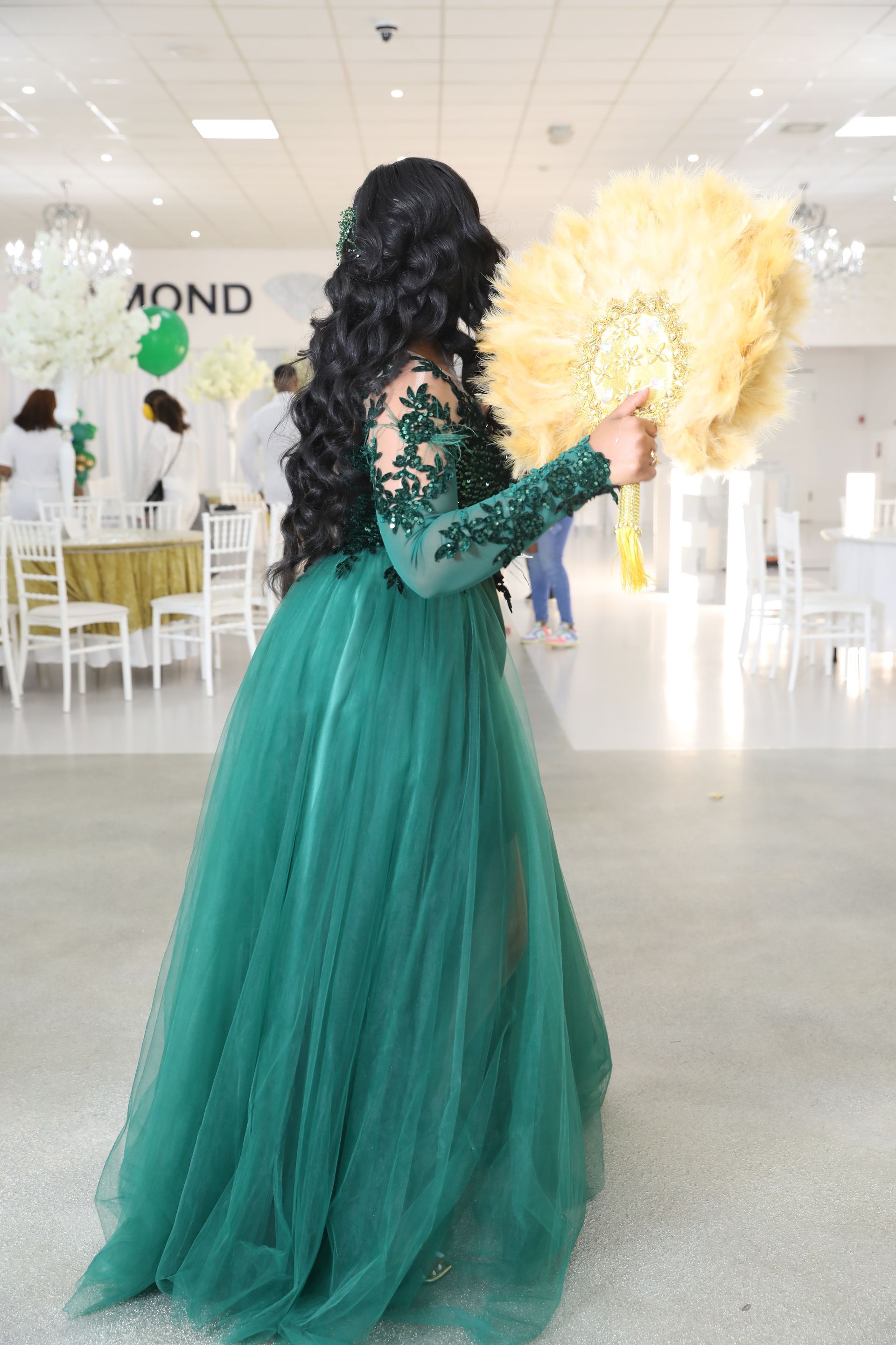 Een vrouw in een groene jurk houdt een grote waaier vast in een goed verlichte evenementenruimte.