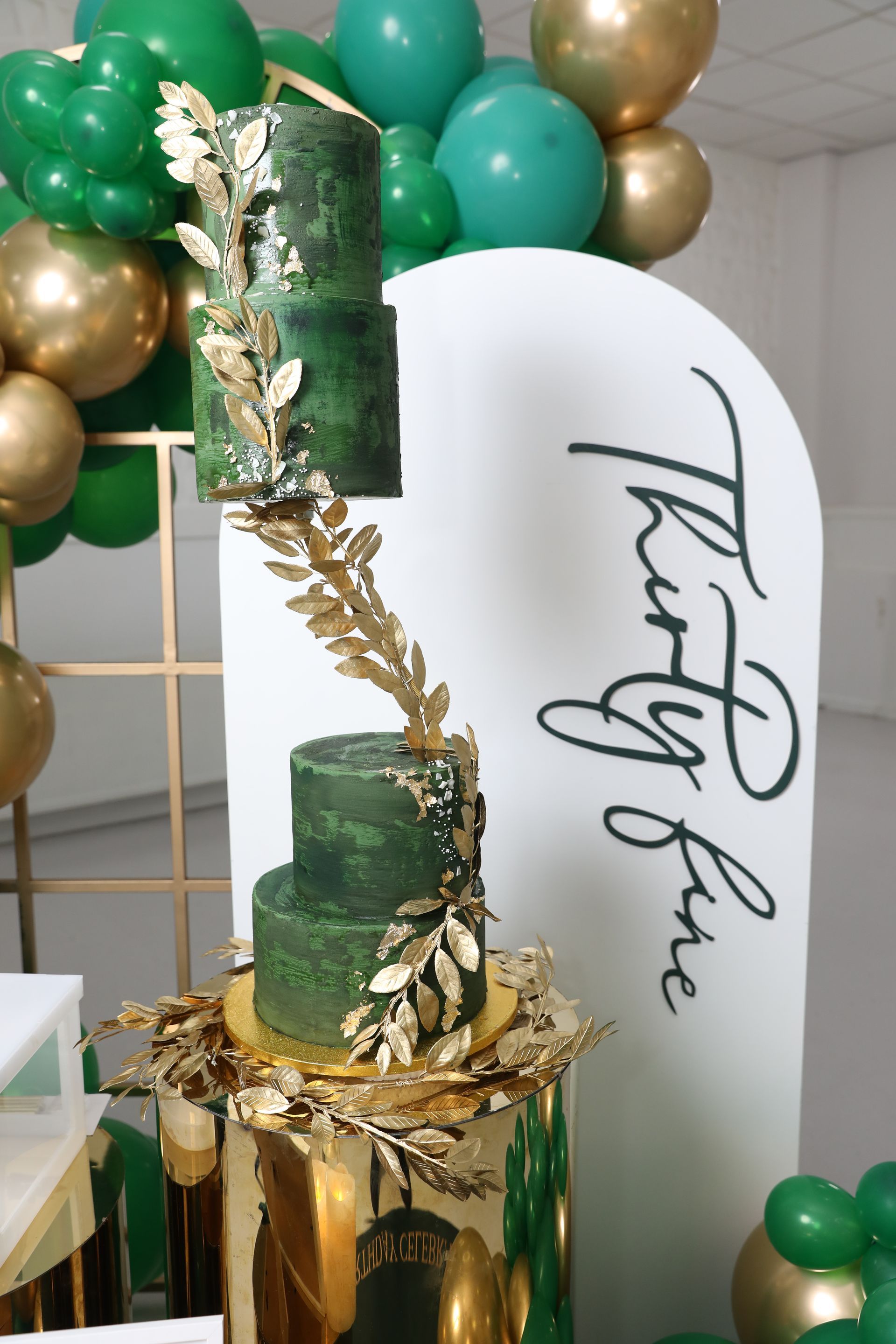 Een groene taart met drie lagen en gouden accenten op een spiegelende standaard, naast een ballonnenboog en een bord met de tekst 
