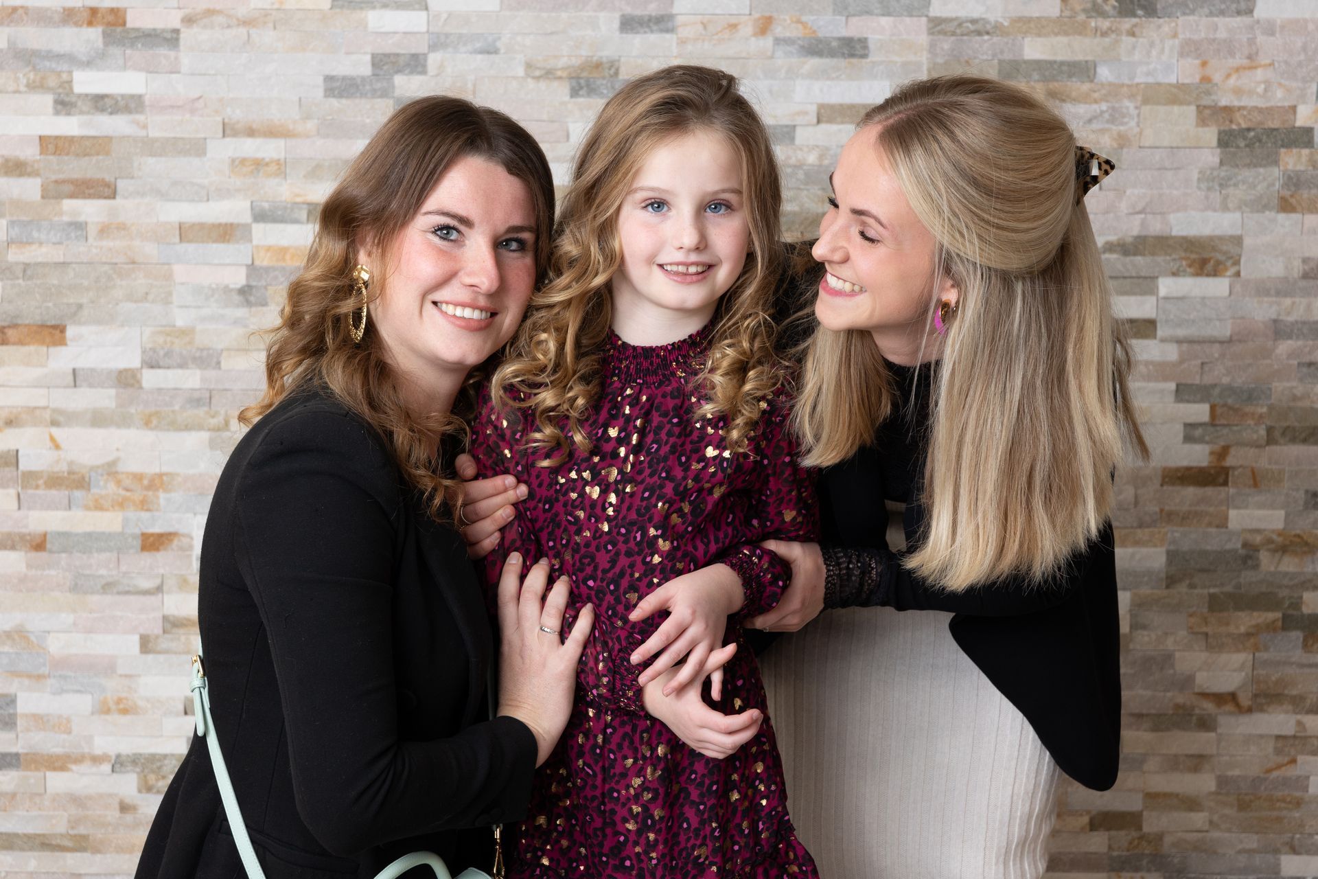 Drie lachende mensen poseren voor een stenen muur. Een kind in een rode jurk staat tussen twee vrouwen in, die elk een arm om haar heen hebben geslagen.