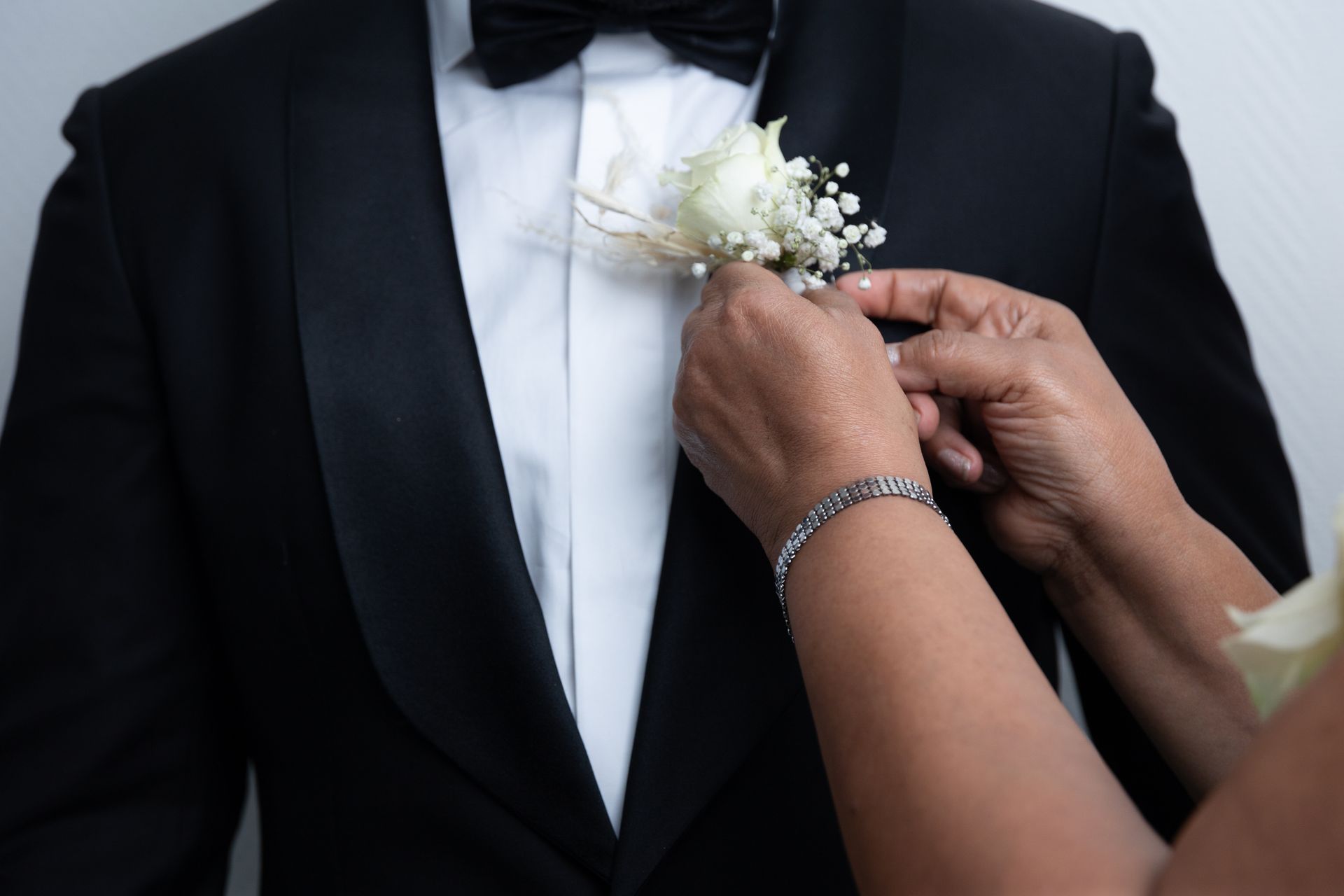 Persoon in smoking krijgt een corsage bijgezet. Handen plaatsen bloemen op de revers; wit overhemd en vlinderdas.