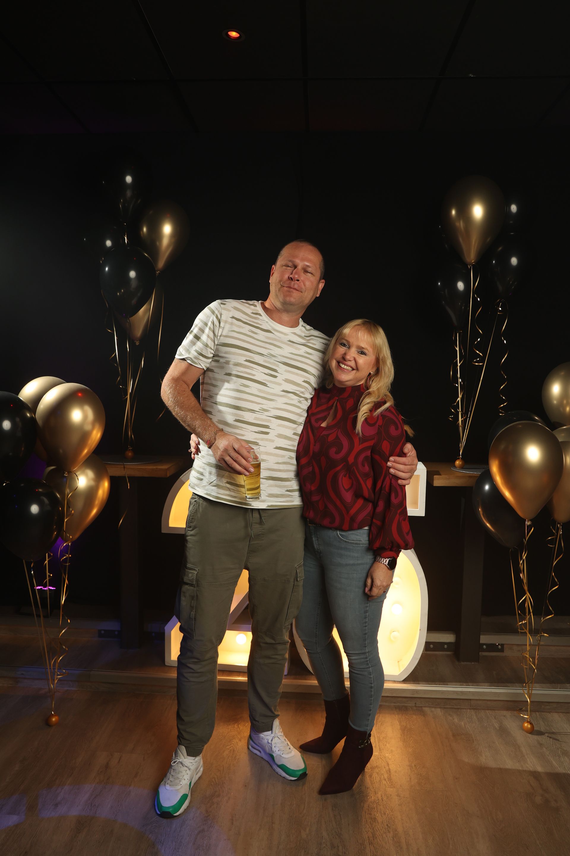 Een man en een vrouw poseren lachend samen op een feestje. Gouden en zwarte ballonnen versieren de donkere achtergrond.