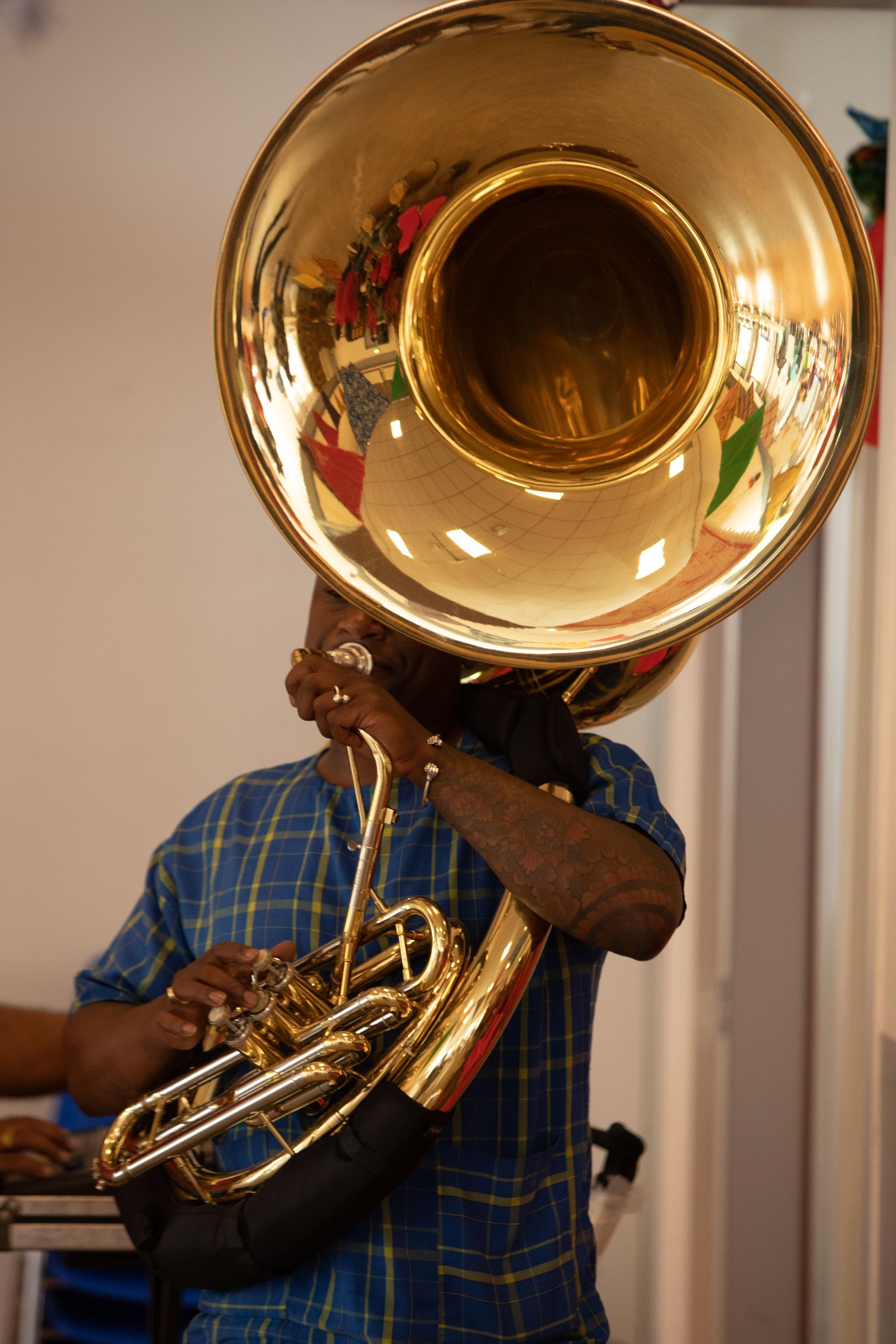 Een man bespeelt een glimmende tuba met een grote klankbeker; hij draagt ​​een blauw geruit overhemd binnenshuis.