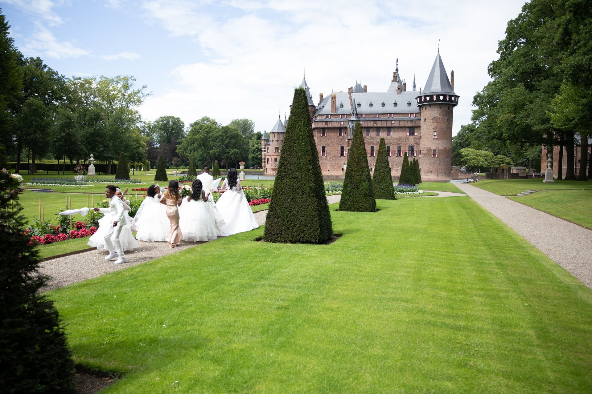 Een groep mensen in trouwjurken loopt op een zonnige dag over een pad richting een kasteel in een tuin.