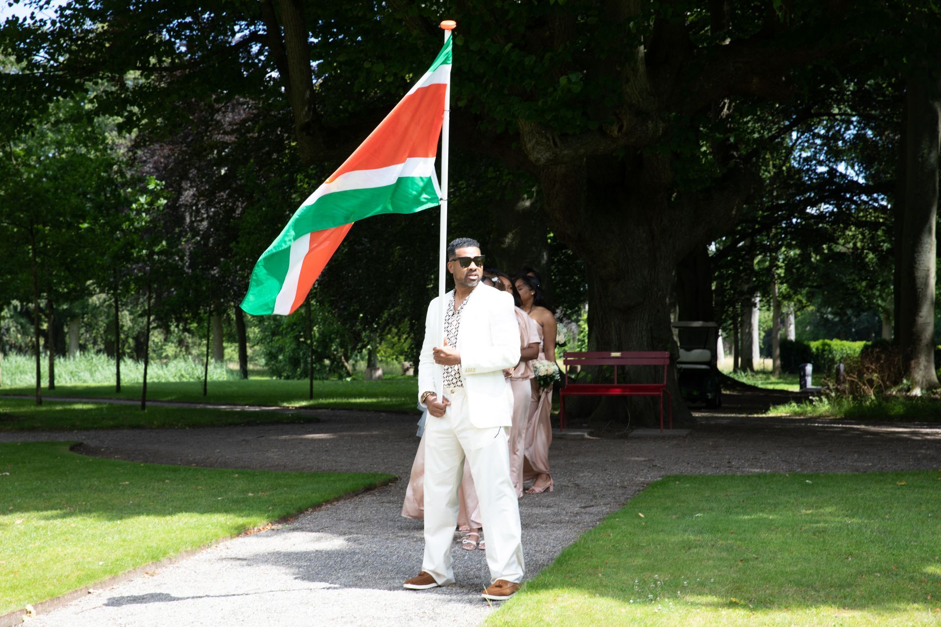 Een man in een wit pak, met een vlag met oranje, witte en groene strepen, loopt met mensen in een park.