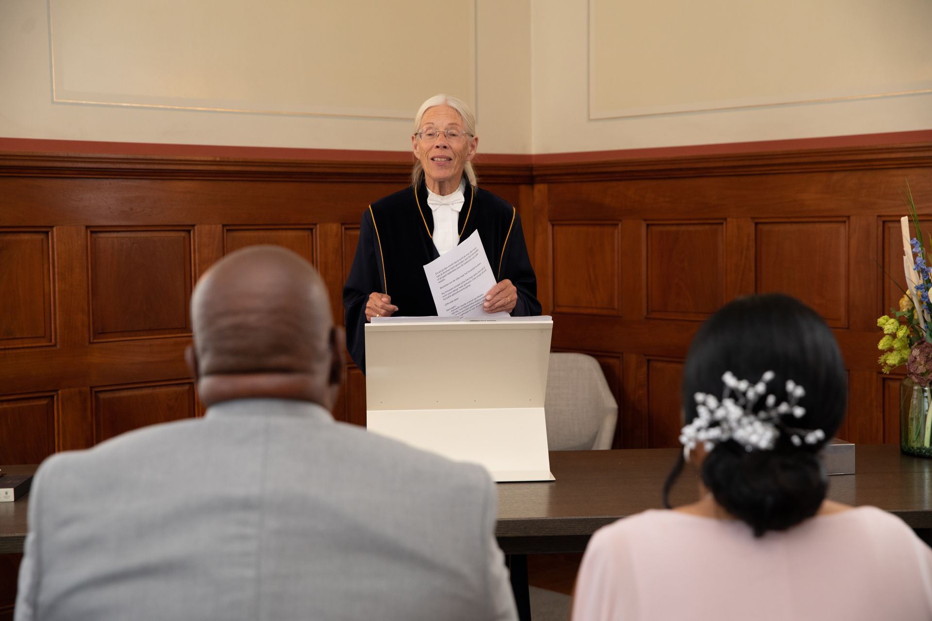 Een persoon met wit haar spreekt op een bruiloft. Een echtpaar zit tegenover de spreker aan een tafel. Op de achtergrond is houten lambrisering te zien.