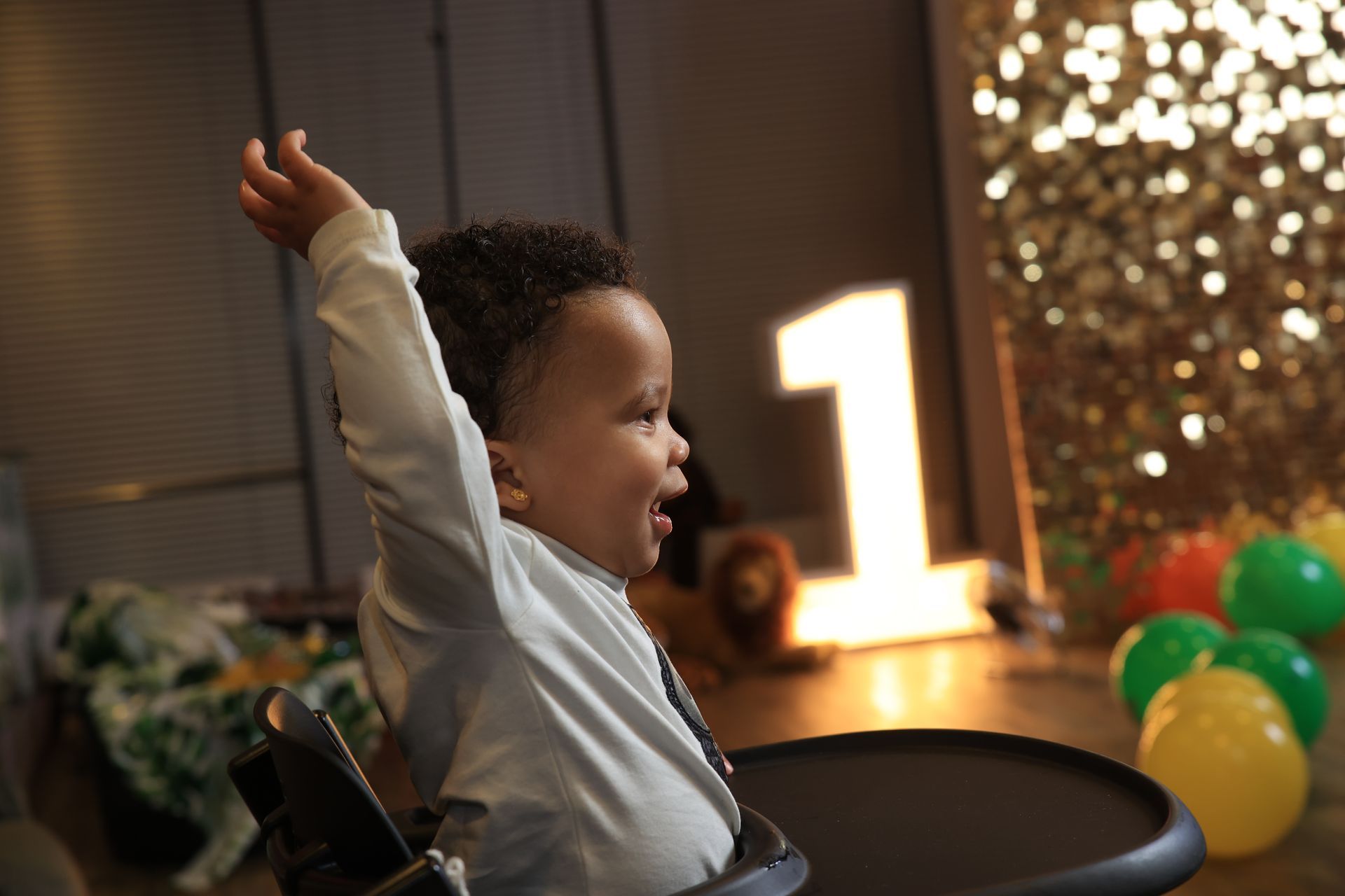 Een lachend kindje in een kinderstoel, met een armpje omhoog, viert zijn eerste verjaardag. Ballonnen en een verlicht cijfer één op de achtergrond.