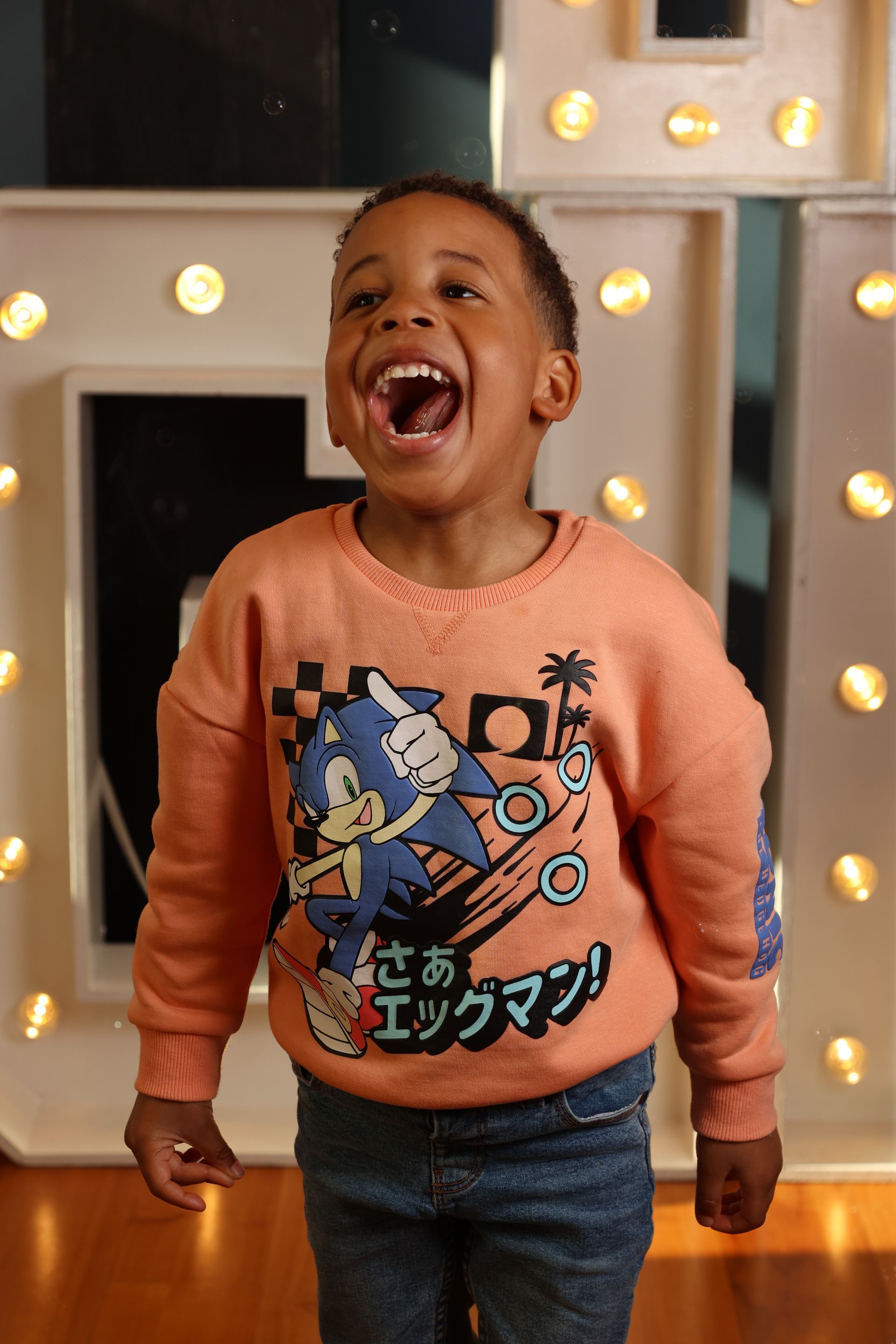Een jong kind lacht, draagt ​​een Sonic the Hedgehog-trui en staat voor verlichte letters.