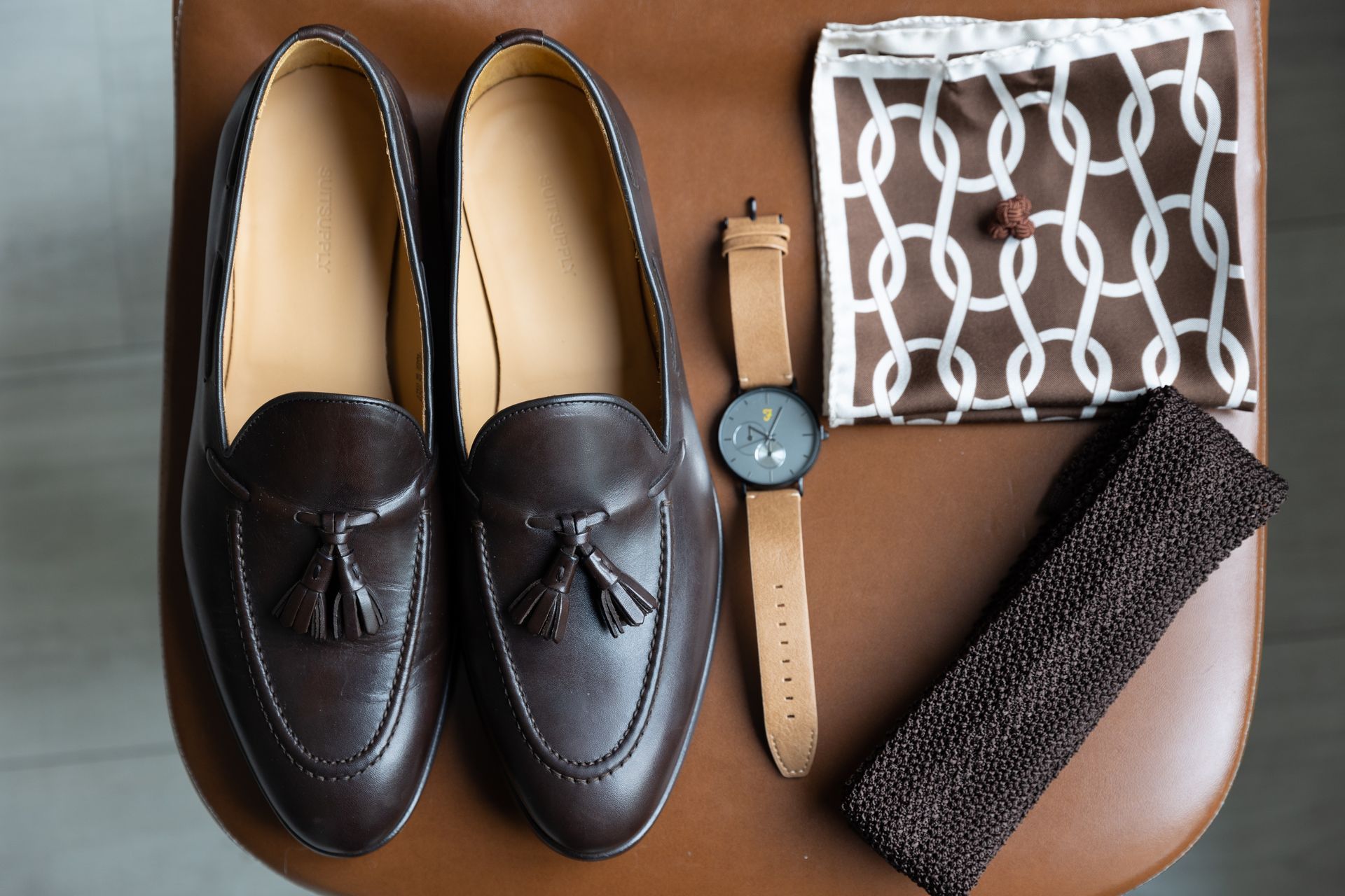 Bruine leren loafers, horloge, poetsborstel en een doek met patroon op een bruine ondergrond.