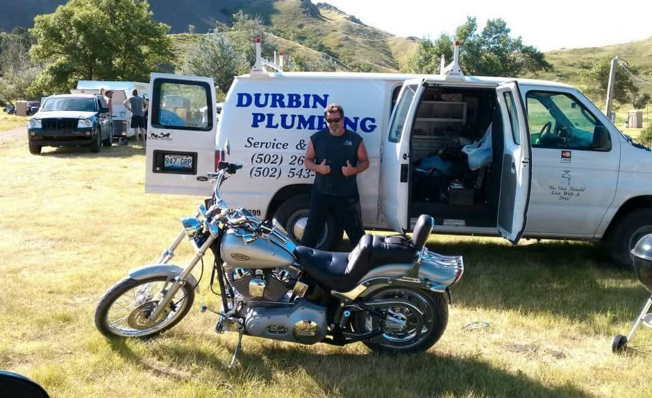 Durbin Plumbing van with worker standing by a black motorcycle in a grassy field.