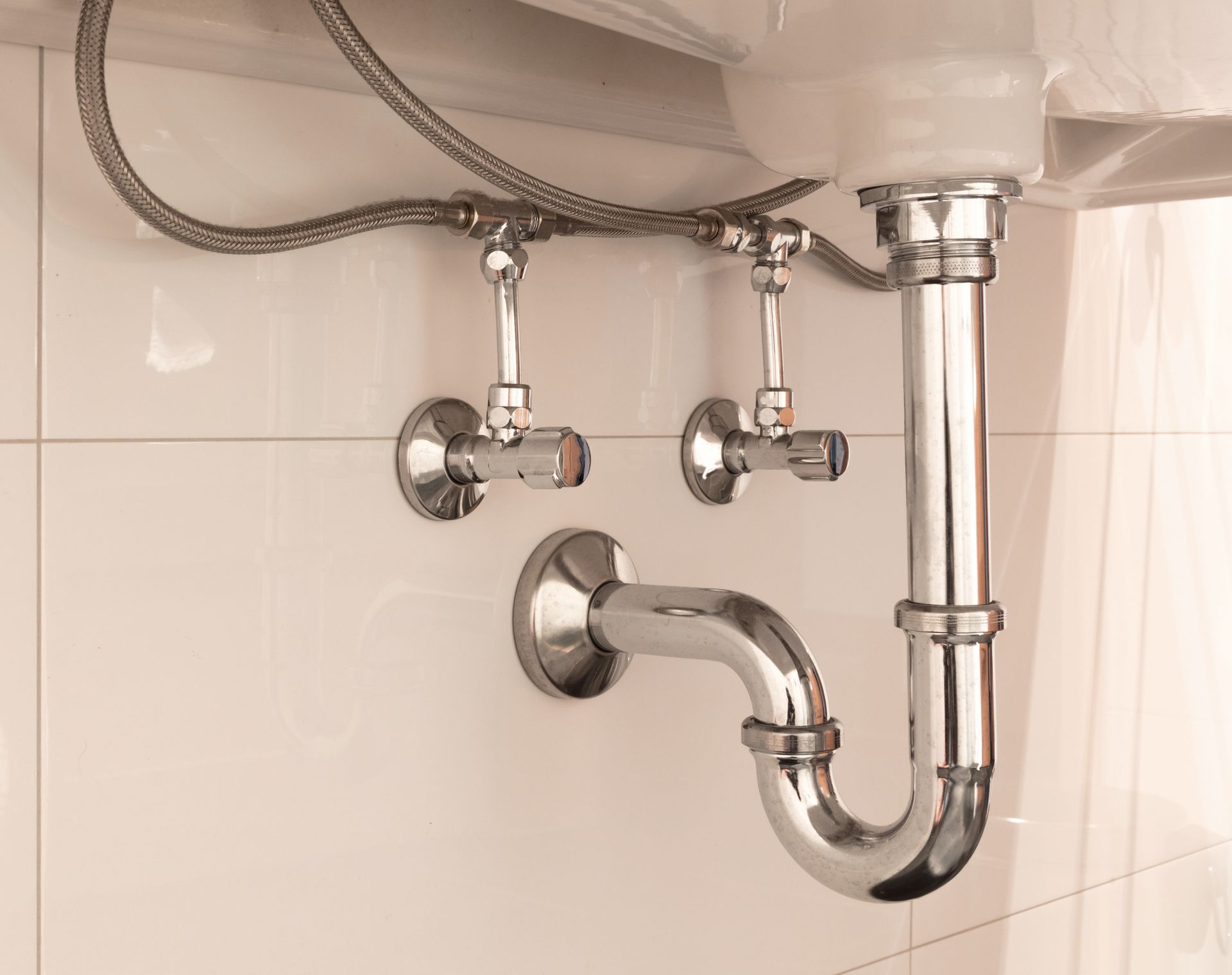 Chrome sink plumbing under a bathroom sink against white tiled wall.