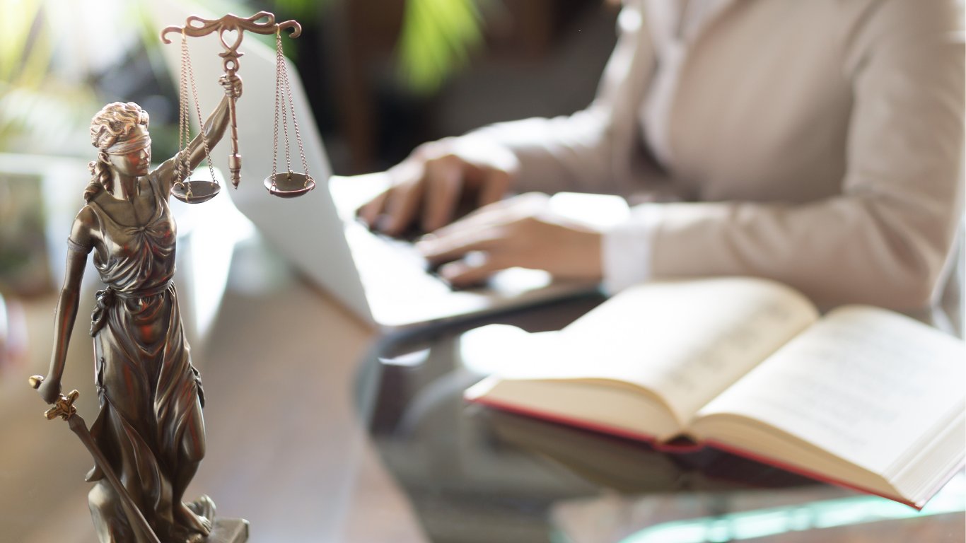 Law book with scales of justice beside a person typing on a laptop at a desk