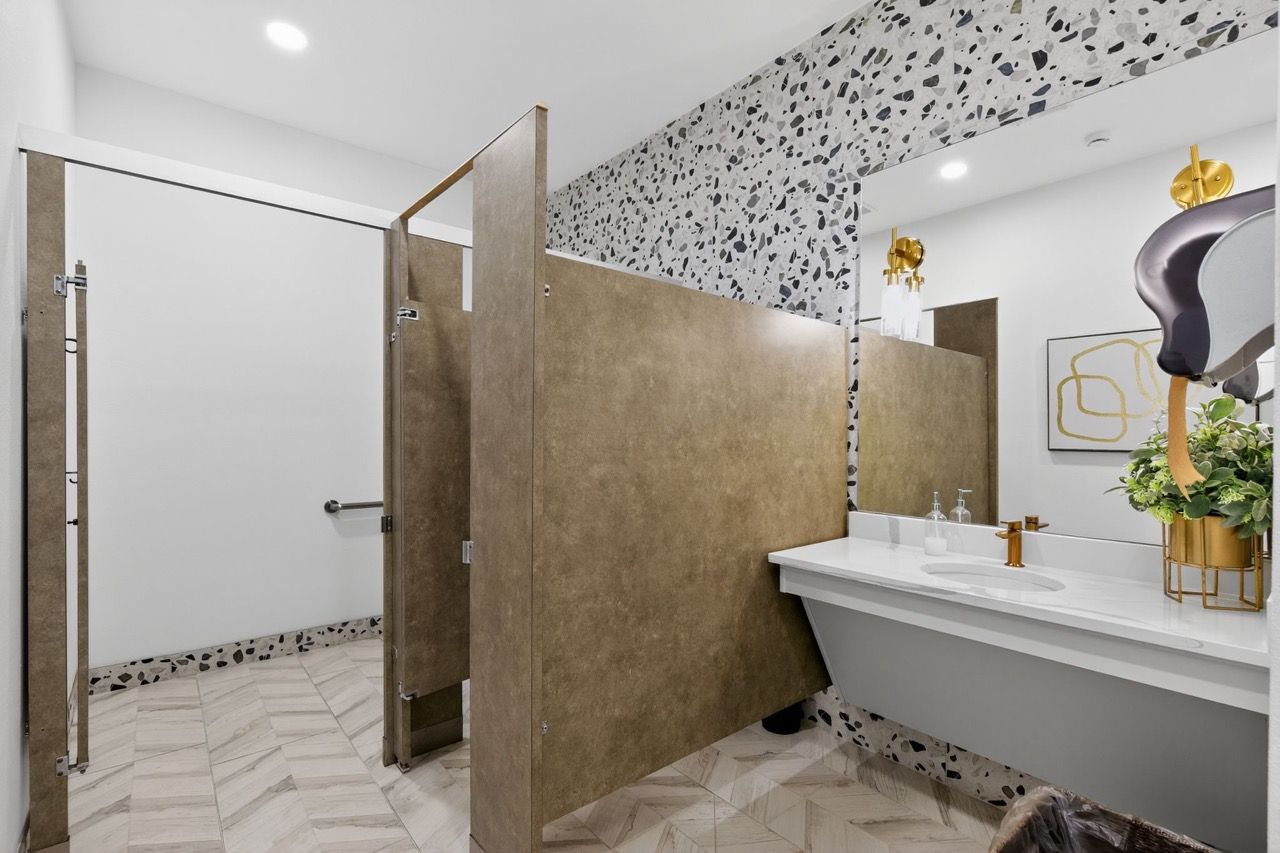 Interior communal bathroom with partition stalls, terrazzo-pattern wall, and a long white vanity with a gold faucet.