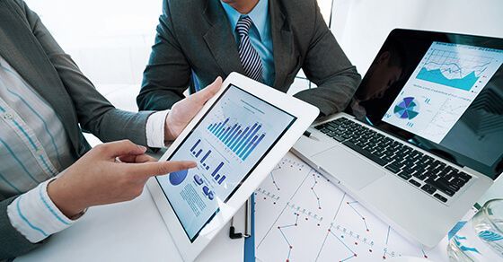 Two businesspeople analyzing charts on a tablet and laptop, collaborating in an office setting.