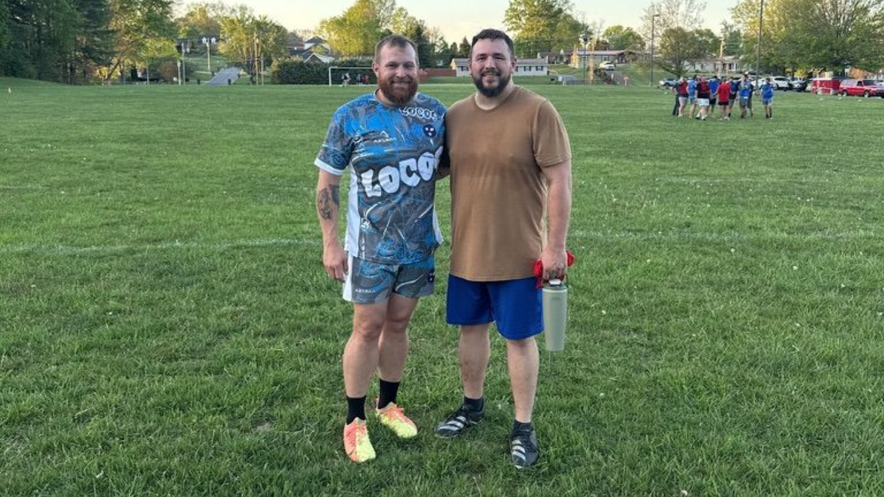 Two men stand side-by-side on a grassy field, smiling at the camera. One wears a patterned jersey and the other a tan shirt.