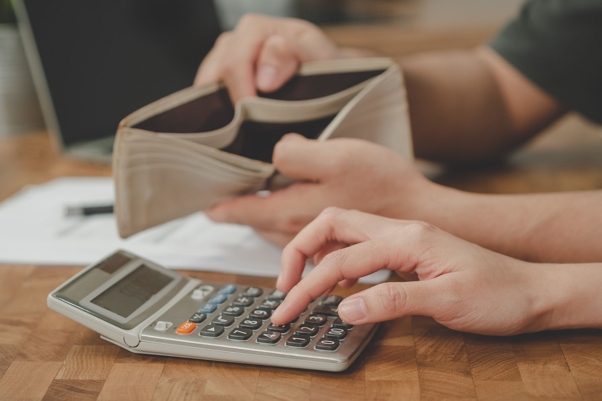 A person is using a calculator while holding an empty wallet.