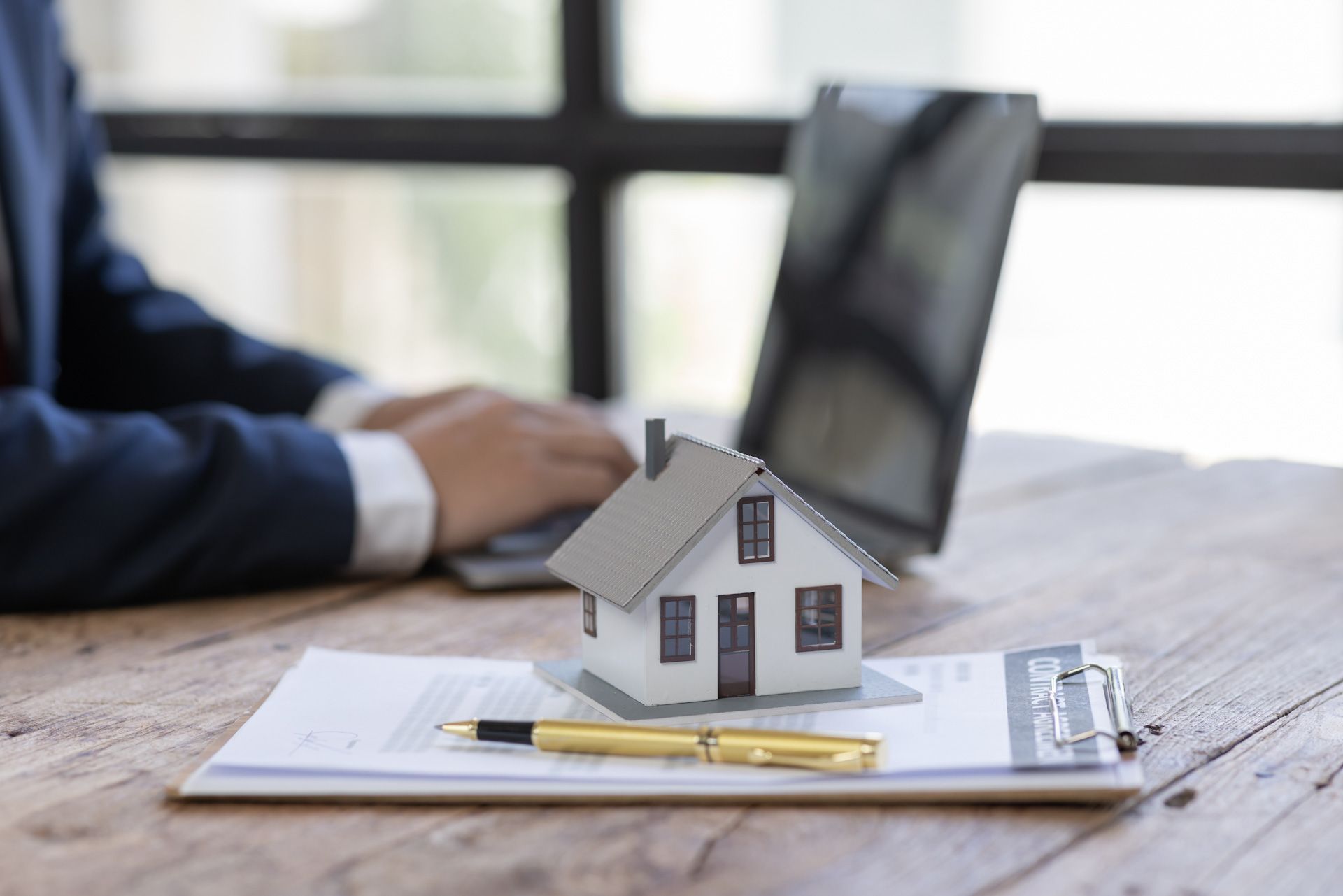 A man is sitting at a desk with a laptop and a model house on top of a clipboard.