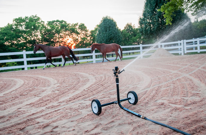 A sprinkler is spraying water on a dirt field with two horses in the background.