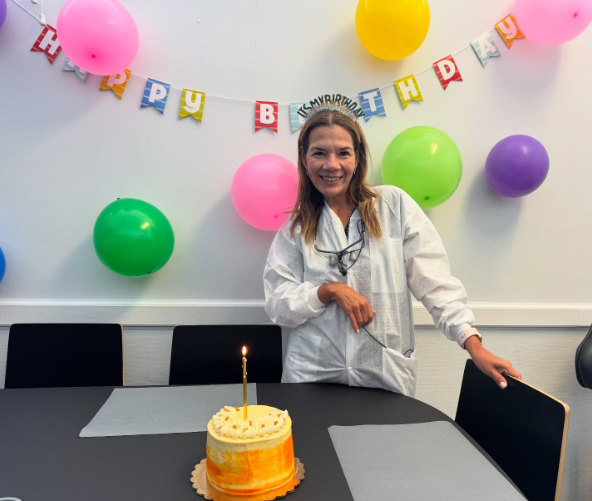 Birthday party setup with balloons, banner, cake, and a smiling person standing by the table