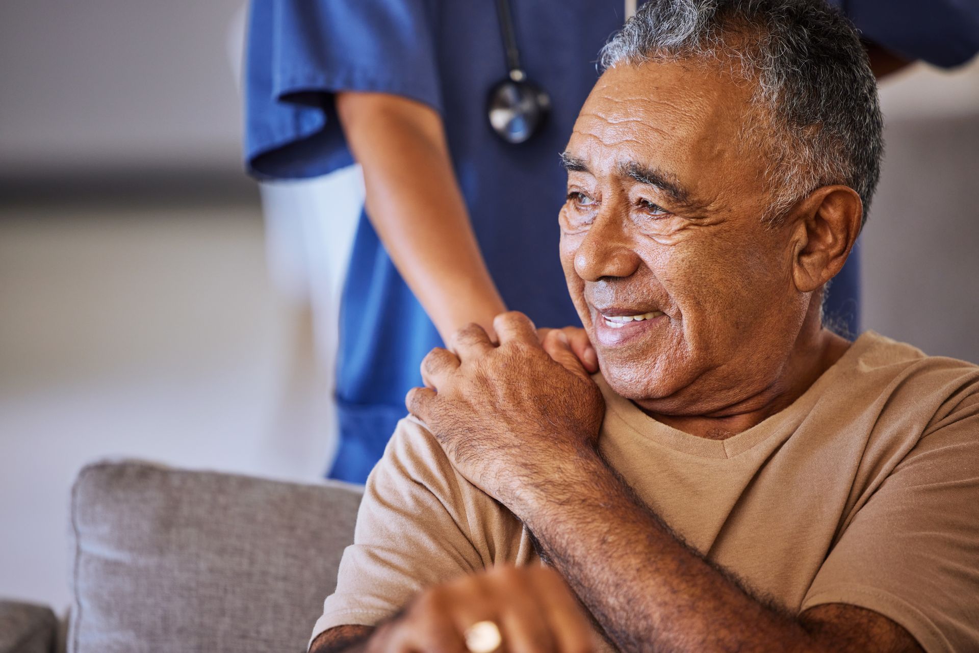 Elderly man smiles, a healthcare worker's hand on his shoulder indoors.