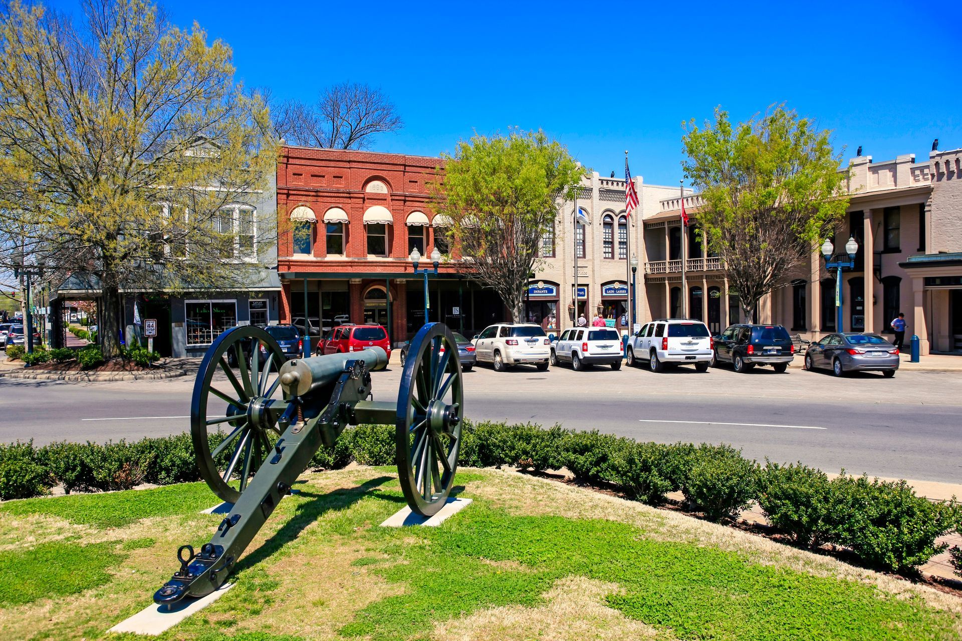 Civil War cannon on a grassy town square with historic storefronts and parked cars behind it