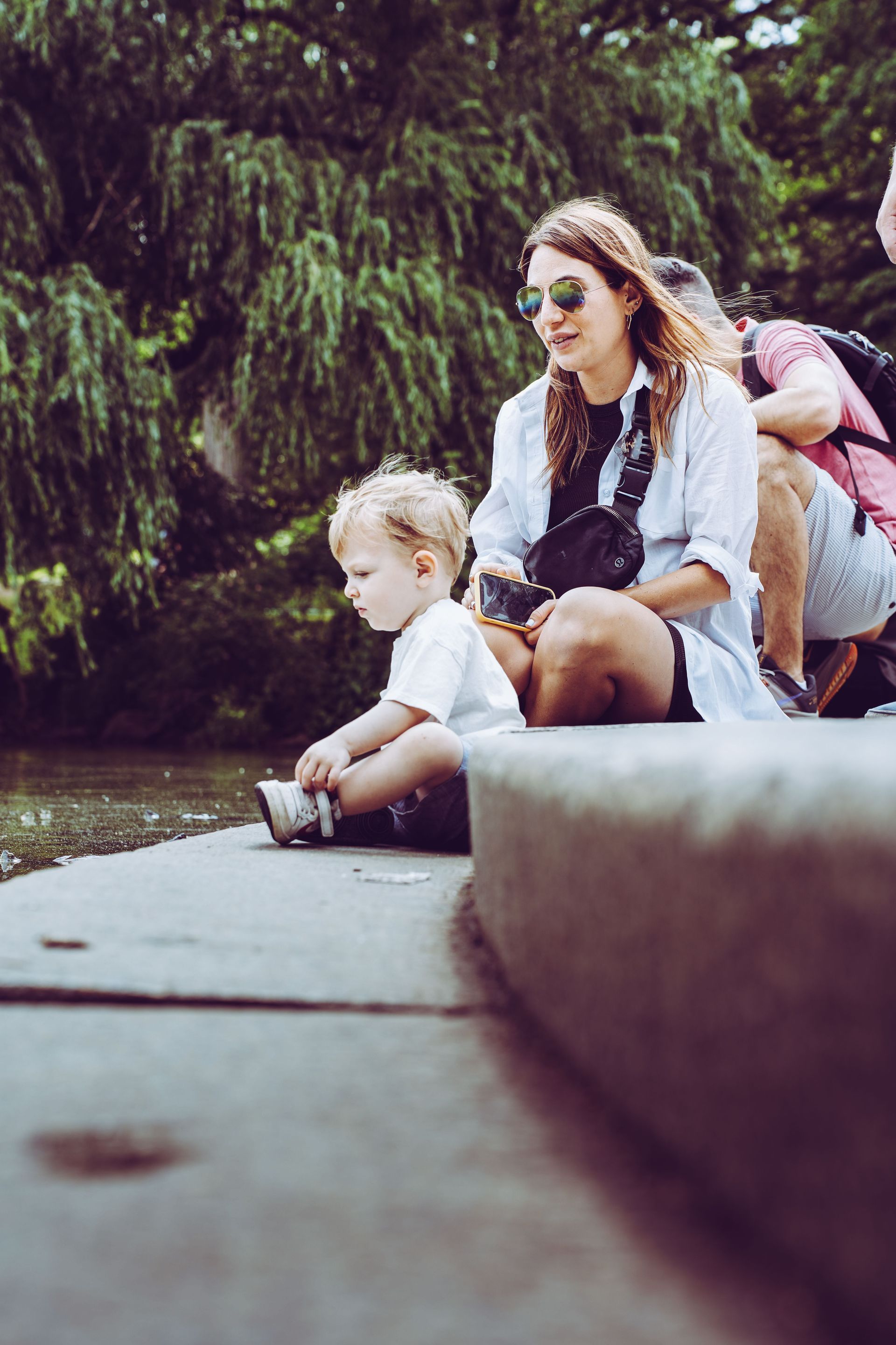 Woman and child sitting by a pond, with another child on her back, surrounded by greenery