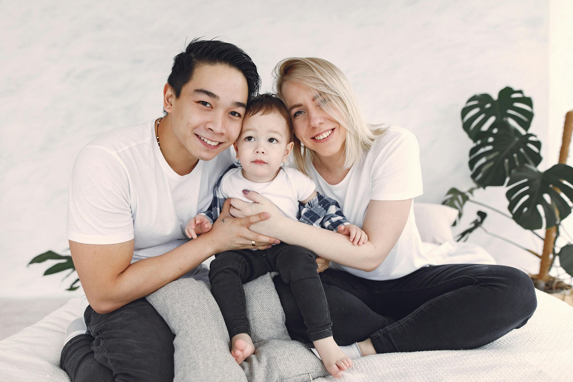 Family of three smiling, posing on a bed with a plant in the background.
