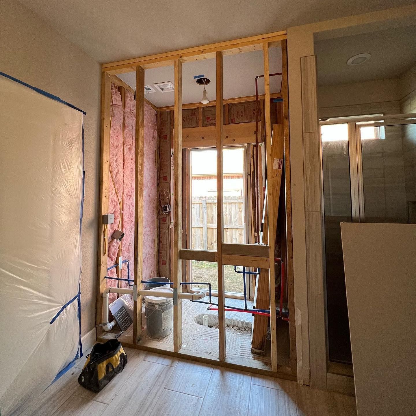 Bathroom under construction, wooden frame, insulation, exposed plumbing, window, and plastic sheeting.