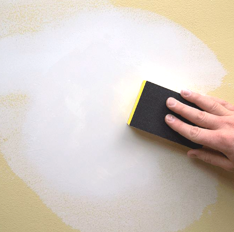 Hand sanding spackling compound on a yellow wall with a sanding block.
