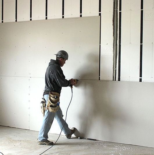 Construction worker installing drywall on a wall with power tool.