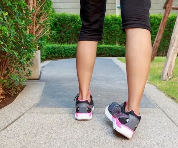 Person walking on a garden path in black leggings and pink-accented sneakers from behind