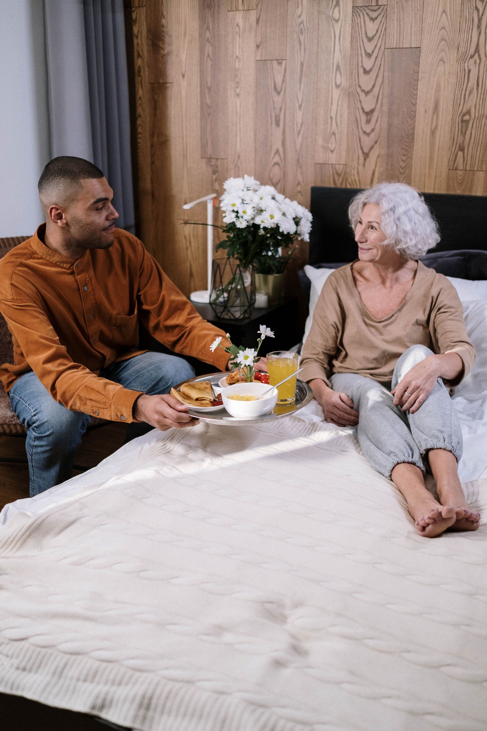 Two people sitting on a bed sharing a meal in a cozy bedroom with flowers nearby